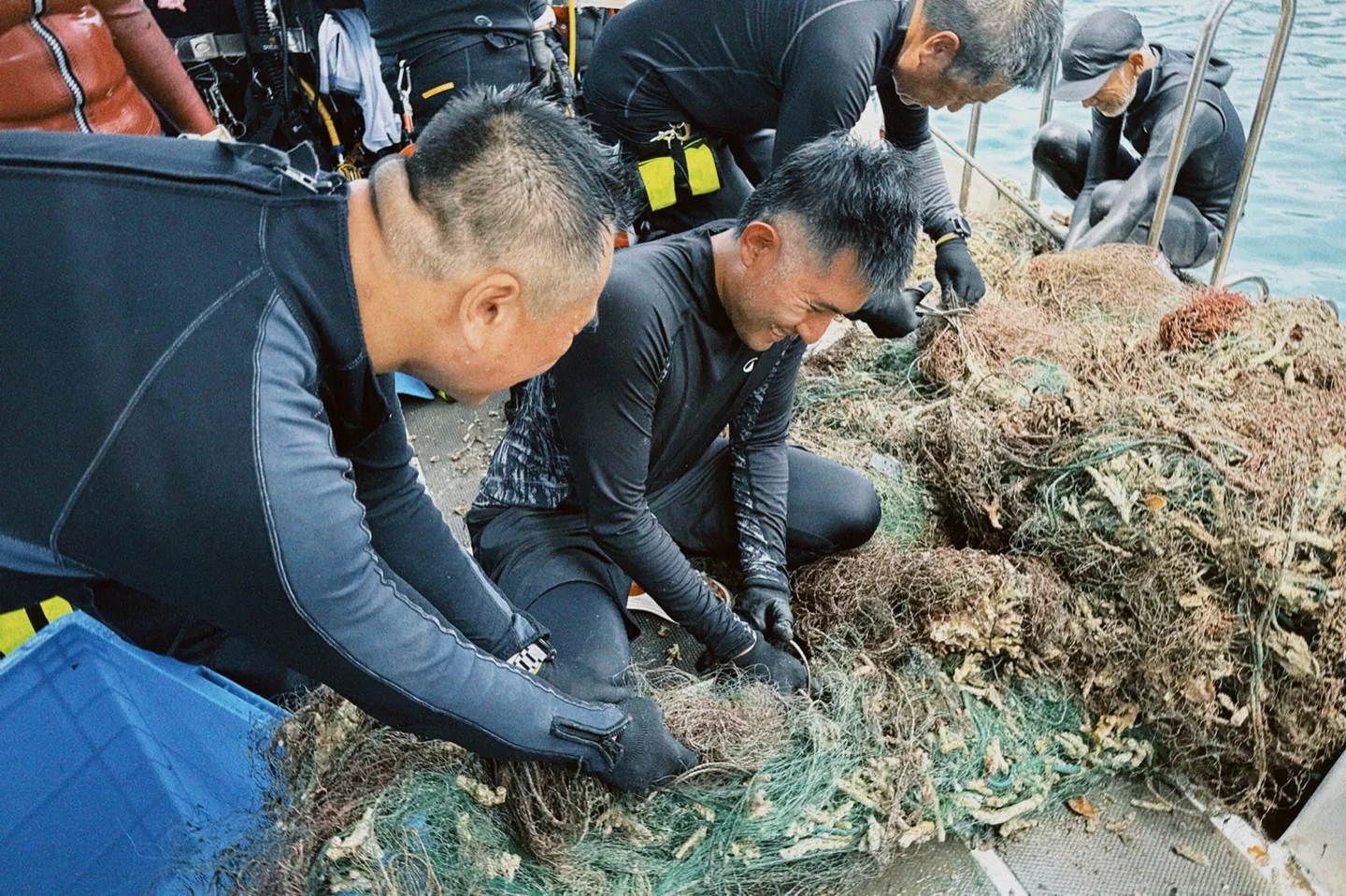 潛水員與工作人員在船上蹲坐一排，合力把剛撈上船的整坨廢棄漁網解開、分類整理